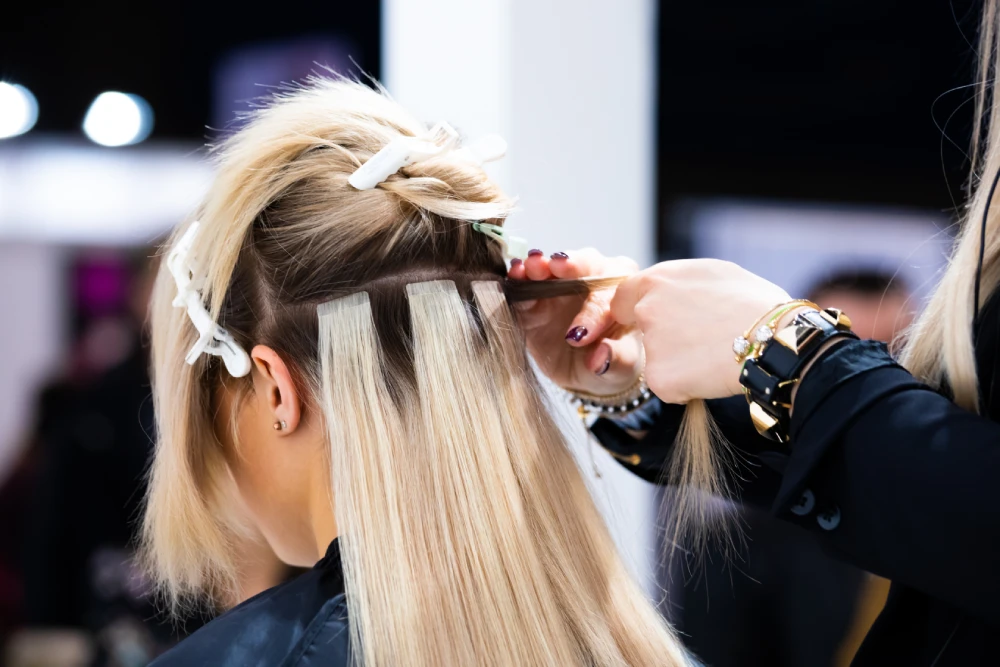 A stylist applies tape-in hair extensions to a client's blonde hair.