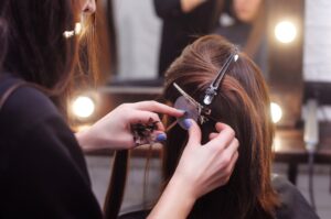 Stylist attaching hair extensions to a woman's parted hair using clips and a tool in a salon.