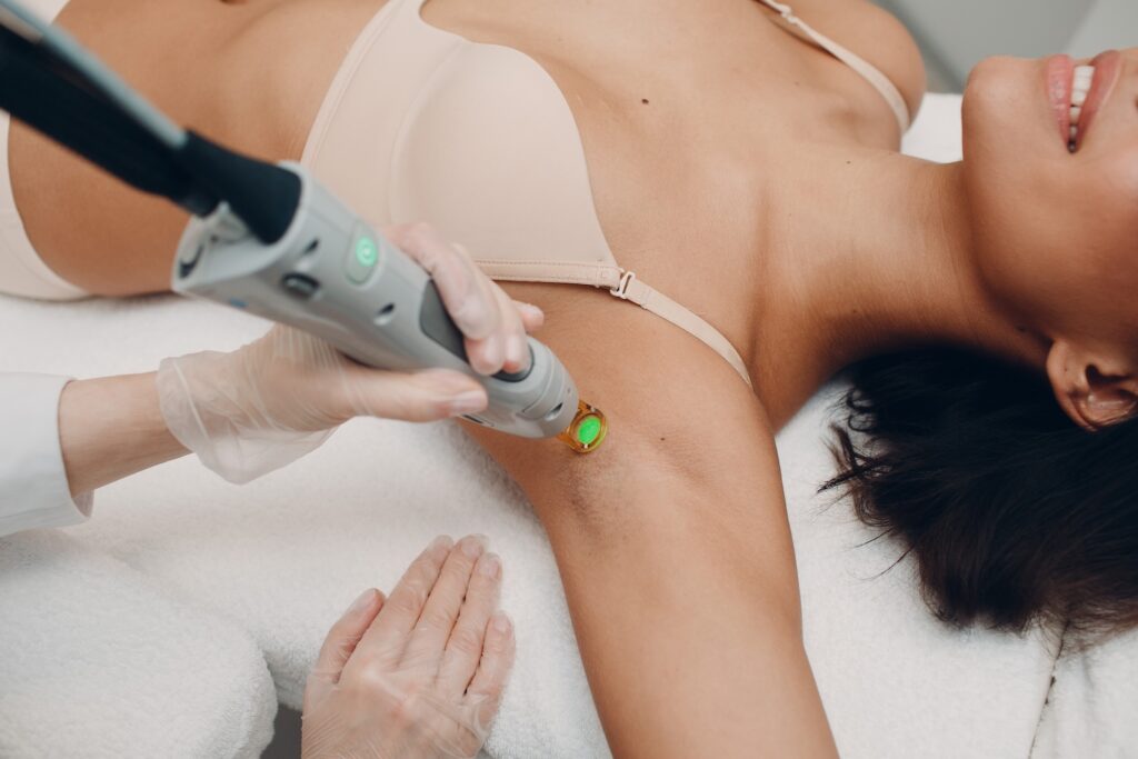 Technician using a handheld laser device on a woman’s underarm while she lies on a treatment bed.