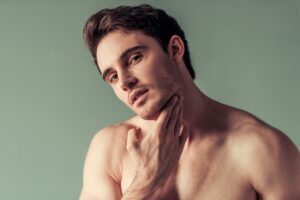 Shirtless man tilting his head and touching his jawline while looking toward the camera.