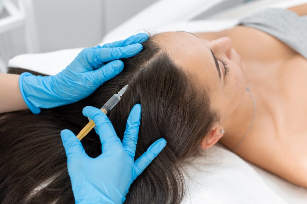 Gloved hands holding a syringe and parting a woman’s hair to inject her scalp as she lies on a treatment bed.