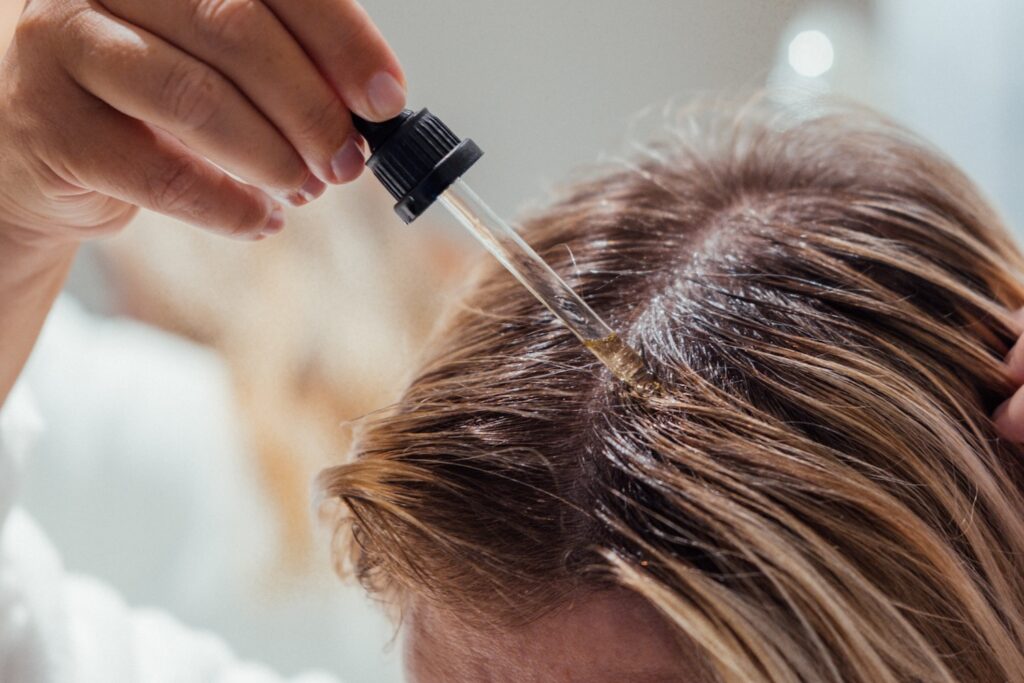 Dropper releasing liquid onto the scalp as hair is parted to expose the application area.