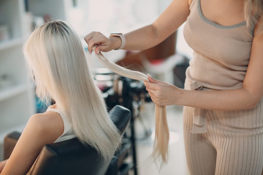 Stylist holding a section of long blonde hair extensions next to a seated woman with light blonde hair.