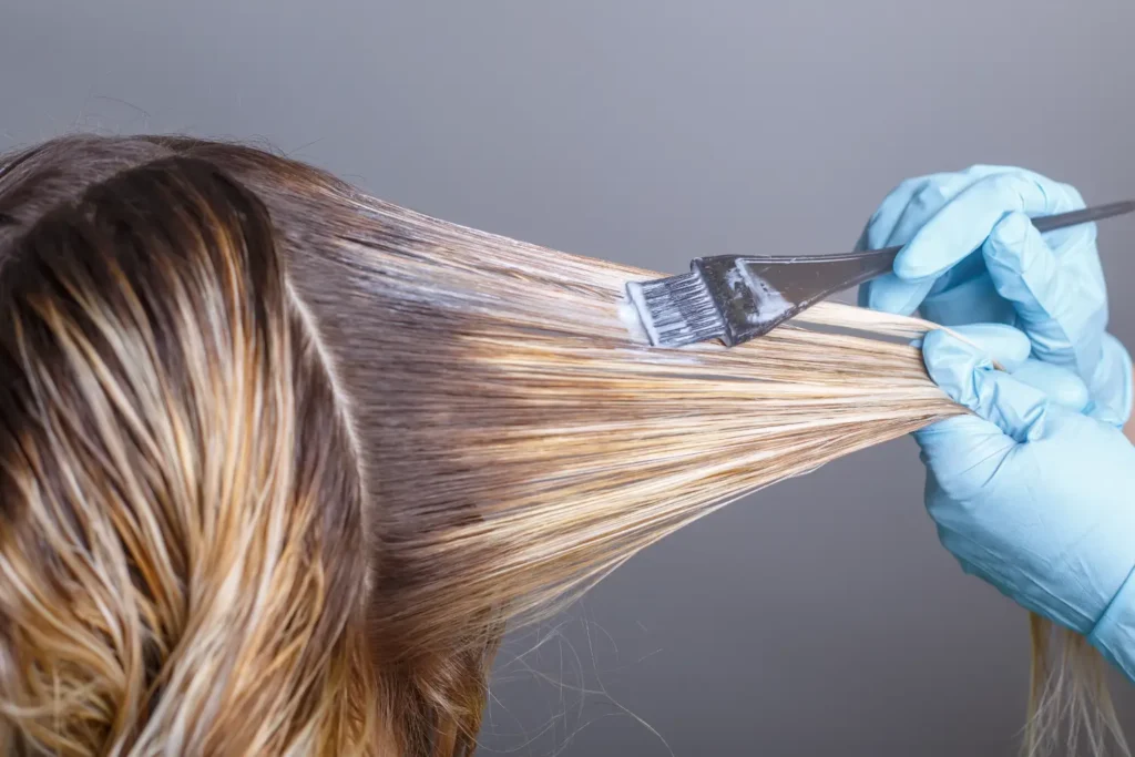 Gloved hands applying hair color to a section of blonde hair with a brush and tint board.