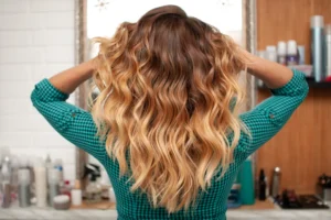 Back view of a person with wavy brown and blonde balayage hair standing in front of a mirror in a salon setting.