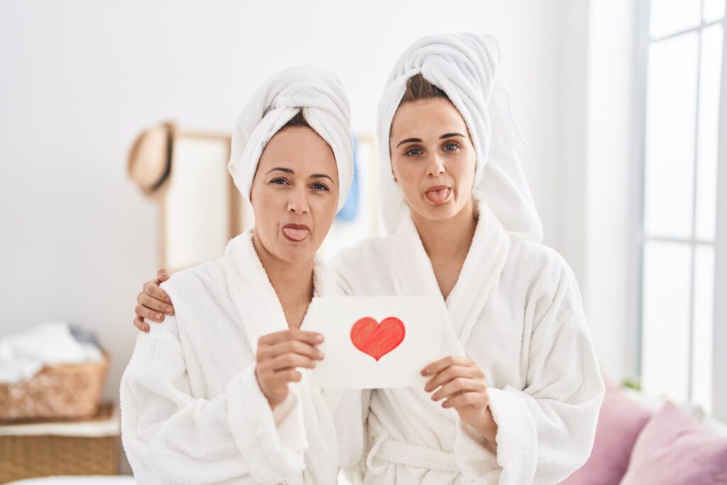 Two women in bathrobes holding card with red heart and sticking tongues out