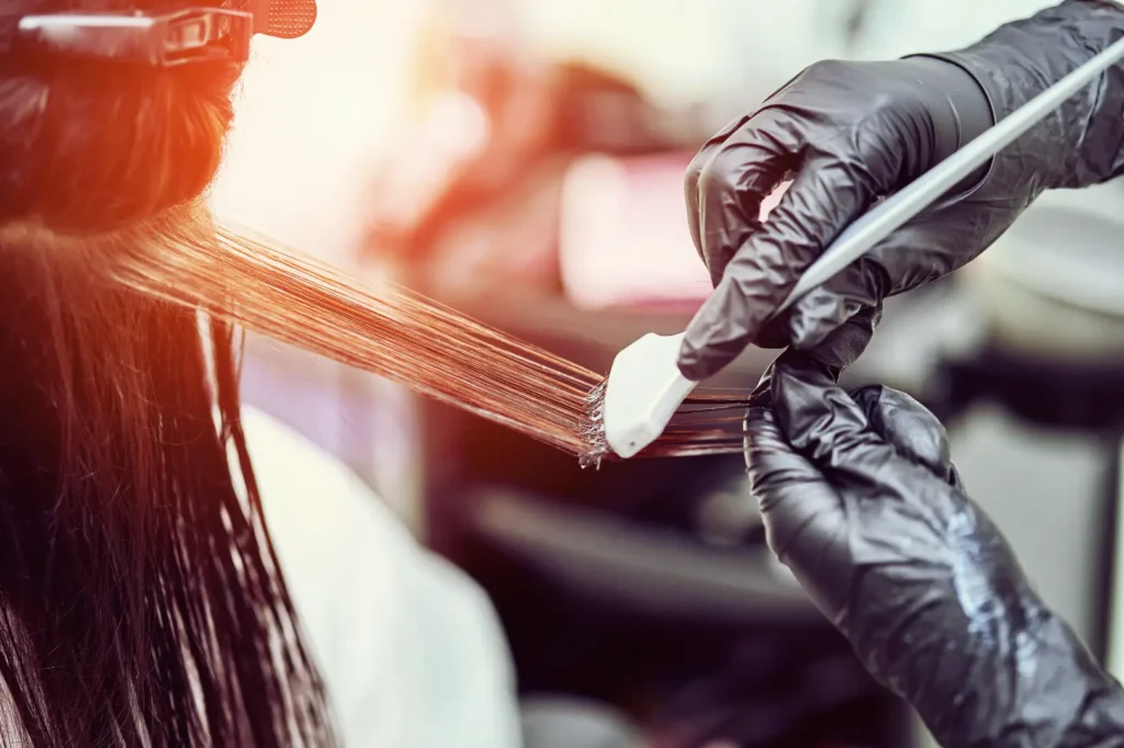 Stylist wearing gloves applies product to a section of highlighted hair with a brush.