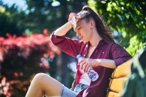 Person sitting outdoors wiping their forehead with a tissue while holding a water bottle on a sunny day.